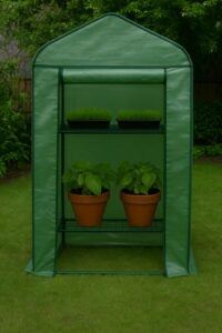 Small portable green greenhouse with metal shelves holding potted plants and seed trays, placed on a grassy backyard lawn with trees in the background.